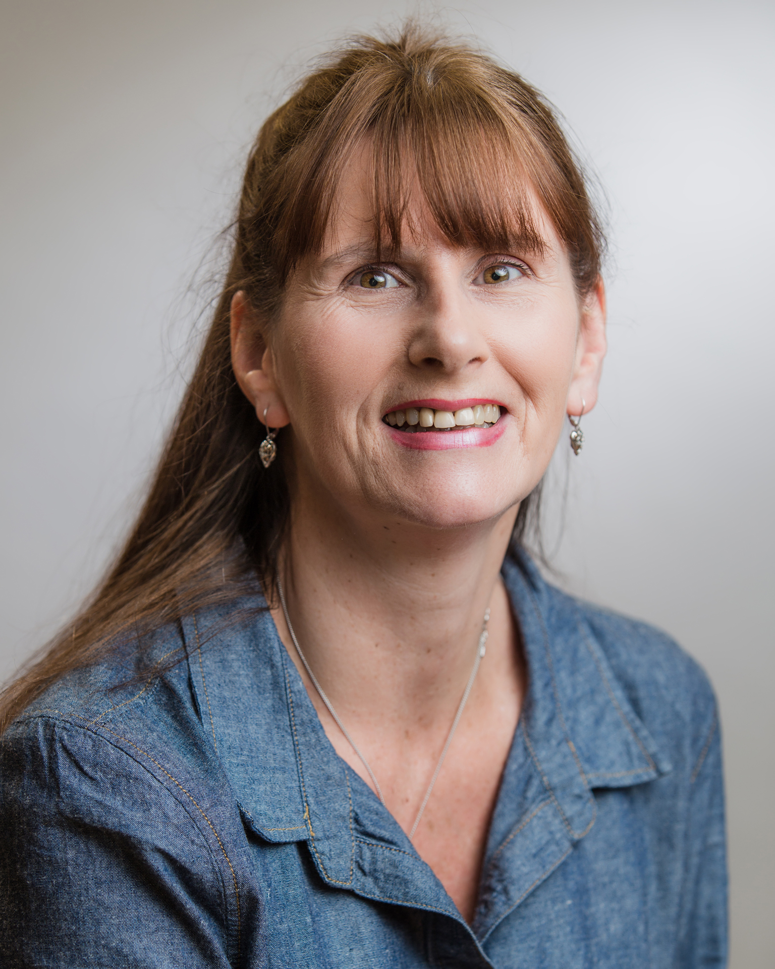 A person with long brown hair and a smart, blue shirt, smiling